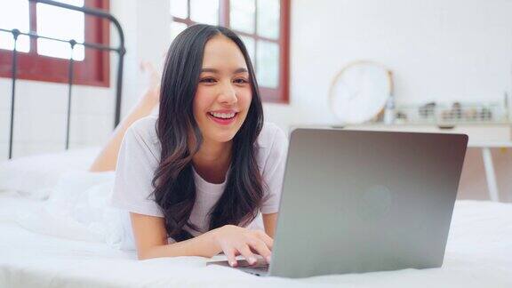 亚洲迷人的女人用笔记本电脑聊天在家里的床上早上快乐的休闲美丽的女性躺在卧室享受假期周末使用笔记本电脑与技术在家里在线交流