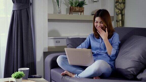 年轻快乐的亚洲妇女工作或浏览互联网与笔记本电脑坐在家里客厅的沙发上