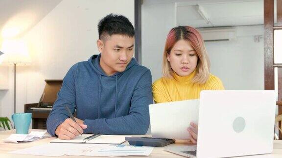 在新房子里一对亚洲夫妇坐在厨房的餐桌旁用纸账单和笔记本电脑计算家庭预算