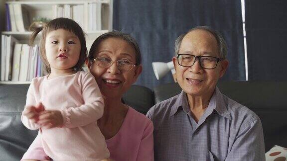 亚洲家庭在客厅里爷爷奶奶和小外甥女孩对着镜头笑着玩耍幸福享受居家生活奶奶抱着可爱可爱的小孩宝宝坐在沙发上