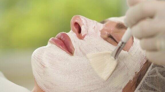 一个女人的脸被白色面具的特写用刷子在脸上涂抹更多的面膜物质