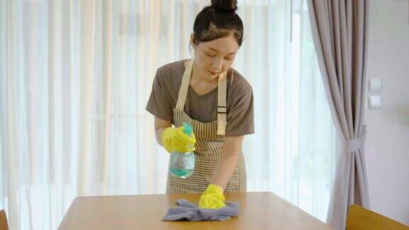 年轻的亚洲妇女戴着手套和围裙抛光或擦拭木制桌子洗涤剂和化学瓶以清除灰尘细菌和消毒家具表面和家务维护