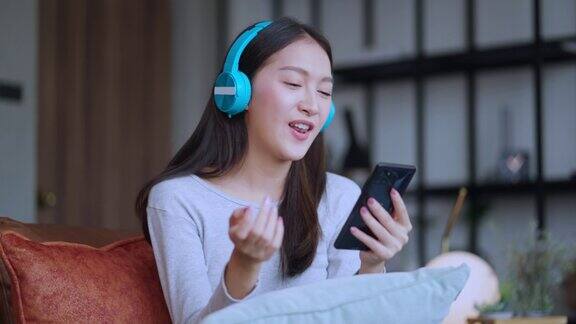 有吸引力的休闲亚洲女性成年女性听音乐耳机积极的情绪坐在沙发沙发旁边的窗户愉快和和平微笑放松亚洲女性使用智能手机在家里白天休闲