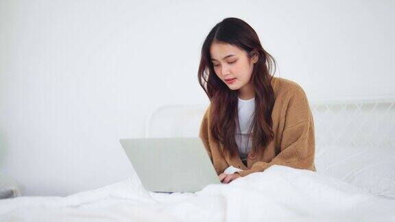 快乐美丽的年轻亚洲女人在电脑上工作坐在家里的床上