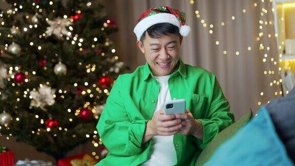 快乐有趣的亚洲人戴着圣诞帽在圣诞新年假期坐在家里使用智能手机快乐阅读短信看搞笑视频浏览聊天手机