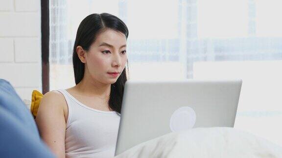 年轻的亚洲女性坐在家里客厅的沙发上用笔记本电脑工作人与科技在家里工作在线教育