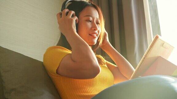 4K亚洲女性坐在沙发上一边听着耳机里的音乐一边用数字平板电脑工作