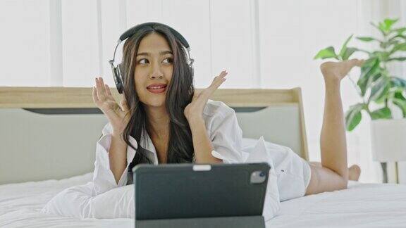 一名年轻的亚洲女性愉快地戴着耳机用笔记本电脑听在线音乐在卧室放松娱乐是生活中的一种小奢侈