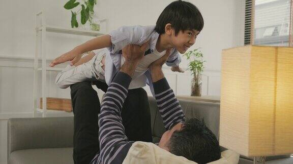亚洲爸爸和儿子在沙发上玩耍双臂像飞机一样伸展