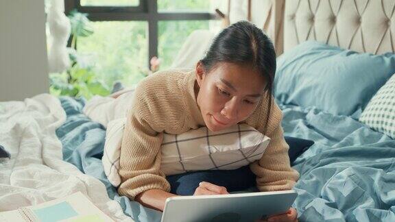 快乐的亚洲女子大学白色的奶油睡衣躺在柔软舒适的床上在家里的卧室里使用数码平板电脑在线学习调整弹性工作制在家办公