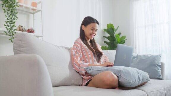 年轻的亚洲女商人在电脑上打字在家工作有魅力的漂亮自由职业女职工坐在沙发上在客厅用笔记本电脑带着幸福和微笑呆在家里