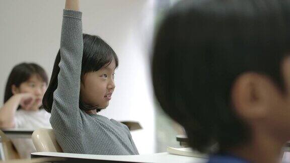 一个亚洲小女孩在教室里举起手臂回答老师的问题幼儿园学前概念