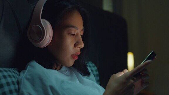 年轻的亚洲女孩沉迷于床上的媒体晚上在卧室里戴着耳机听音乐音频流媒体广播用手机在线播客