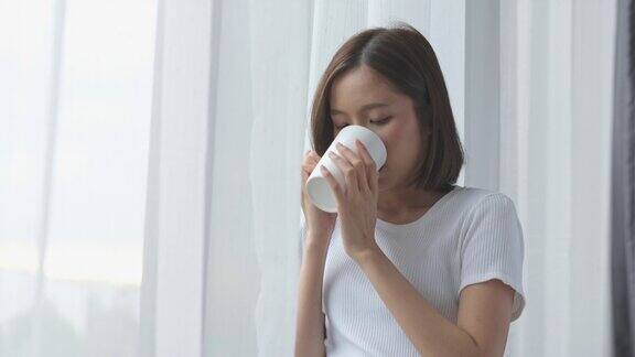 年轻的亚洲女人站在窗前拿着一杯咖啡在家里