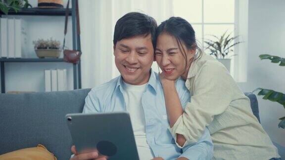 年轻的亚洲夫妇与休闲坐在沙发上使用平板电脑自拍在线呼叫挥手手势与家人和朋友在家里的客厅