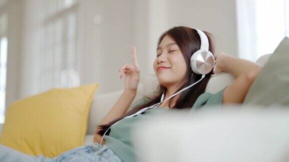 年轻的亚洲女人在家里客厅的沙发上听音乐快乐的亚洲女性使用手机戴着耳机坐在沙发上