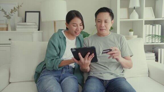 幸福的亚洲夫妇一起坐在家里客厅的沙发上玩电子平板设备购物使用信用卡在线支付生活方式花时间在家里