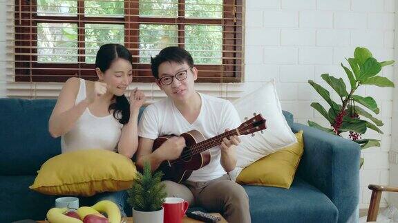 两个年轻的亚洲夫妇家庭放松在沙发上弹吉他音乐和唱歌亚洲生活方式的家庭概念