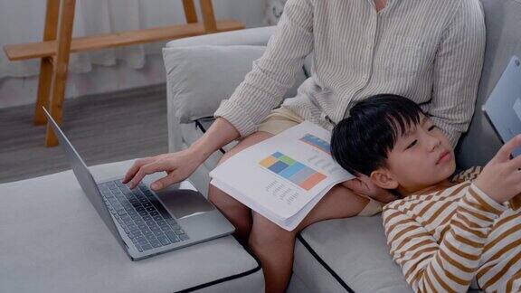 4K中年亚洲单身妈妈在笔记本电脑上工作躺在沙发上儿子躺在她腿上妈妈玩平板电脑男孩玩平板电脑困睡在妈妈的腿上