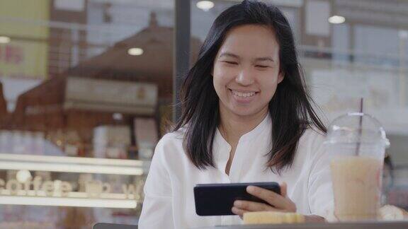 年轻的亚洲女性坐在购物中心的咖啡店前一边吃零食和咖啡一边用智能手机看视频