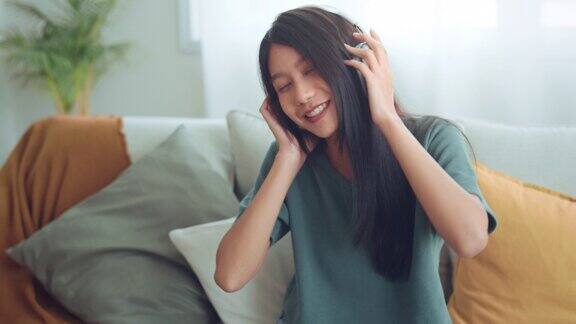 快乐的亚洲年轻女性戴着无线耳机在智能手机上听音乐坐在客厅的沙发上女性喜欢听寒冷的音乐声音感觉没有压力在家里