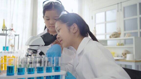 在学校的科学实验室里两个穿着大衣的可爱的亚洲女孩正目不转睛地盯着试管里的混色实验和显微镜概念教育科学实验化学