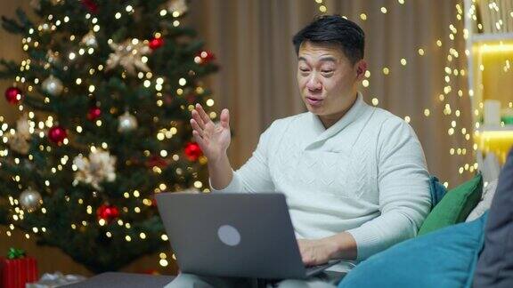 英俊的亚洲男子远程视频通话圣诞夜用笔记本电脑坐在家里的沙发上快乐