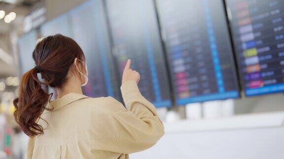 年轻的亚洲女子戴着口罩拿着护照和登机牌在国际机场看着航班信息板检查她的航班