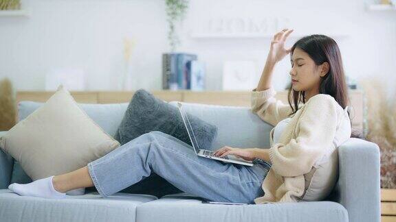 年轻的亚洲女子坐在家里的沙发上使用笔记本电脑和手机