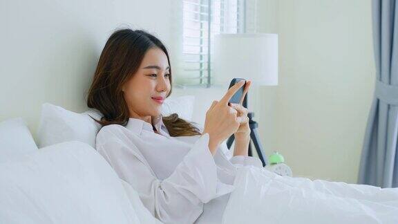 亚洲美女早上在家里的床上用手机聊天快乐随意的漂亮女性躺在卧室享受假日周末在家里用智能手机和科技交流