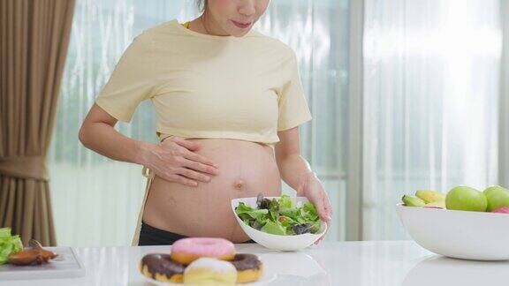 亚洲年轻孕妇选择吃蔬菜沙拉而不是甜甜圈迷人美丽的怀孕女孩站在厨房的室内感到快乐享受吃蔬菜健康食品的健康在家