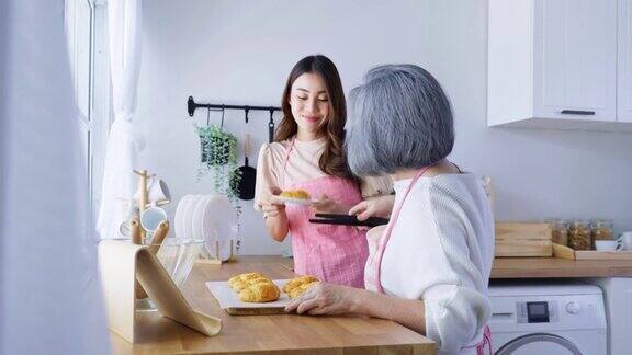 亚洲可爱的家庭年轻的女儿看着老妈妈在厨房做饭漂亮女性享受与年长的妈妈在家里餐桌上烤牛角面包的闲暇时光活动的关系