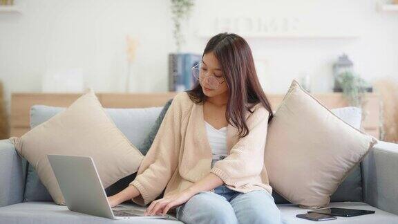 年轻的亚洲女子坐在家里的沙发上使用笔记本电脑