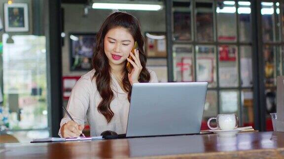 美丽迷人的亚洲女人工作与笔记本电脑微笑并在智能手机与客户交谈以获得想法和要求的成功工作外的办公室在咖啡店商业概念暖色调