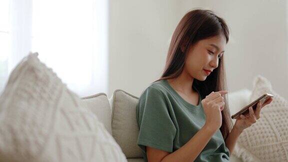 快乐的年轻亚洲女性放松在舒适的沙发上在家里用智能手机发短信微笑的女孩使用手机聊天浏览无线互联网上的小工具从家里网上购物