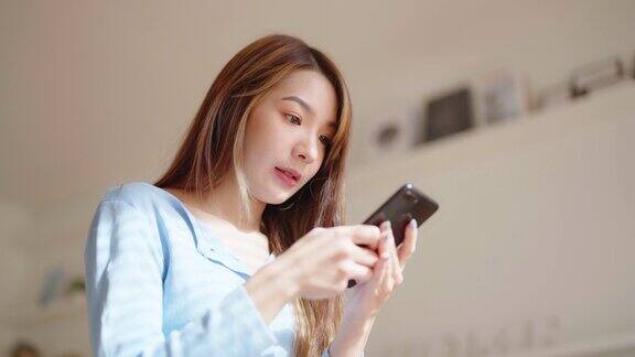 快乐的年轻亚洲女子放松在舒适的沙发上在家里发短信在手机上微笑的女孩使用手机聊天浏览无线互联网上的小工具从家里网上购物