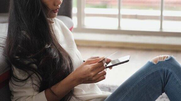 年轻的亚洲女性用智能手机和信用卡支付网上购物坐在家里的沙发上数字时代的生活方式