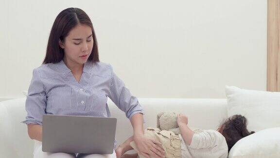 亚洲女性母亲在客厅笔记本电脑上工作孩子女孩睡在沙发上