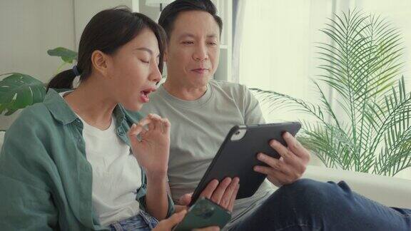 幸福的亚洲夫妇坐在家里客厅的沙发上一起玩智能手机和平板电脑网上购物生活方式花时间在家里