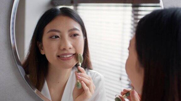 美丽的年轻亚洲妇女看着镜子使用绿宝石玉压路机