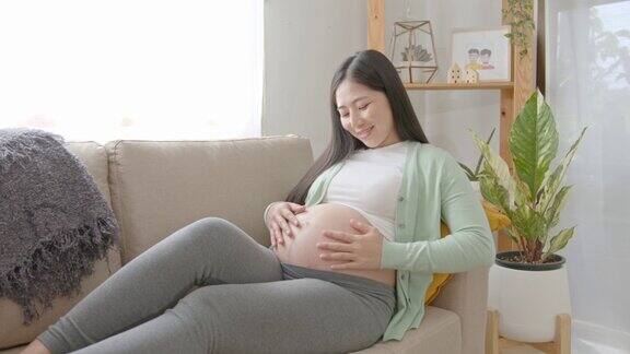 年轻的亚洲孕妇手爱抚腹部坐在沙发上在家里怀孕产妇准备和预期