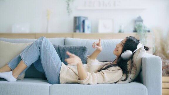 年轻的亚洲女子在家里客厅的沙发上听音乐快乐亚洲女性使用手机戴耳机躺在沙发上