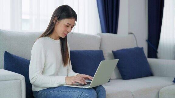 年轻的亚洲女性坐在家里的沙发上使用笔记本电脑