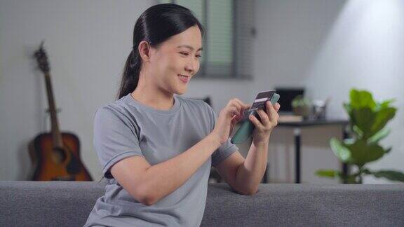 亚洲女性坐在沙发上用智能手机和信用卡在家里网购