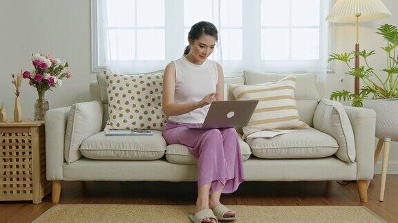 年轻的亚洲女商人在沙发上用笔记本电脑工作在家工作