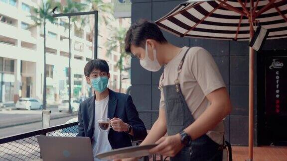 亚洲自由职业者戴着外科口罩在咖啡馆用笔记本电脑打字时喝咖啡