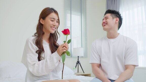 亚洲青年惊喜送玫瑰给美丽的女友迷人浪漫的新婚男女坐在床上一起享受庆祝周年纪念日和情人节