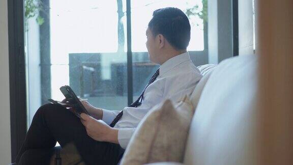 资深亚洲金融商人坐在办公室的沙发上一边拿着数字平板电脑一边写着货币股票投资的金融策略一边思考和规划商业金融计划一边看向外面
