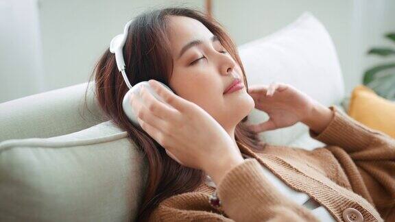 宁静的亚洲女人放松在舒适的沙发上闭着眼睛戴着耳机在客厅女性喜欢听寒冷的音乐声音声音在家里感觉没有压力