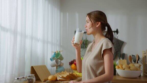 亚洲年轻美丽的女人在家里的厨房喝一杯牛奶迷人的女性望着窗户早上醒来后端着一杯牛奶在家里保健品